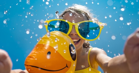 young girl wearing a bright swimsuit and sunglasses holding a duck-shaped flotation device under water and smiling